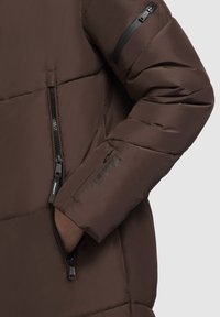 Jachetă puffer maro cu un exterior lucios, cusături orizontale, buzunare cu fermoar și un buzunar pe mânecă cu fermoar, având accente negre.