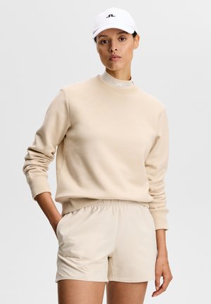 Kvinde iført en beige sweatshirt, matchende shorts, en hvid højhalset undertrøje og en hvid baseballkasket med hånden i lommen.