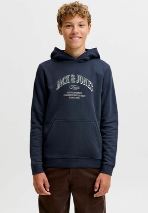 Adolescent aux cheveux bouclés portant un sweat à capuche bleu marine Jack & Jones et un pantalon marron, souriant et debout sur un fond uni.