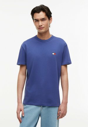 Giovane uomo che indossa una maglietta blu a maniche corte con un piccolo logo sul petto e jeans azzurri, in piedi davanti a uno sfondo bianco.