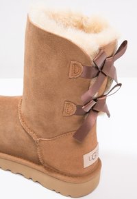 Tan suède enkellaars met zachte crème kleurige bontvoering, decoratieve bruine lintstrikjes aan de achterkant en een lichtbruine rubberen zool. Logo-label zichtbaar.