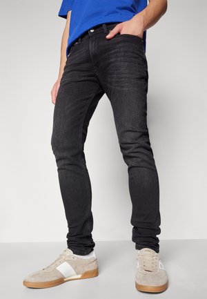 Homme portant un jean noir ajusté, une chemise bleue et des baskets beiges, debout avec une main dans la poche sur un fond uni.