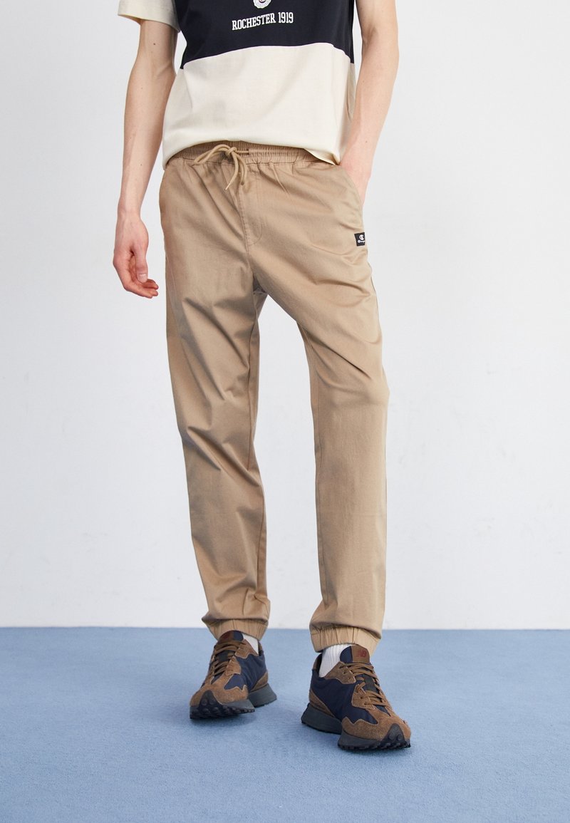 Beige joggerbyxor med elastisk midja, sidofickor och cuffed ankle-kant. Bärs med mörka sneakers med beige detaljer.