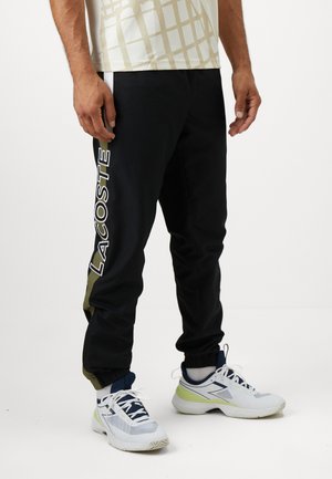 Pantaloni sport negri cu logo lateral alb, accente verde măsliniu, manșete elastice și un design elegant. Asortați cu adidași albi și gri.