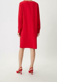 Vestido largo rojo de manga larga con corte holgado, puños abotonados y escote redondeado. Combinado con tacones de aguja rojos con correas en los tobillos.