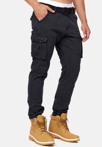 Pantalones cargo negros con múltiples bolsillos, cinturón ajustable y un ajuste entallado. Combinados con botas de encaje marrones, con suela resistente.