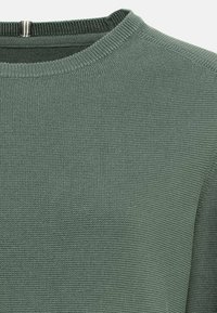 Gros plan sur un pull en tricot vert montrant l'encolure ras du cou côtelée et le motif de tricot texturé.