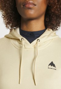 Krämfärgad hoodie med dragsko, prydd med en svart bergslogotyp på vänster sida och en mjuk, texturerad tyg.