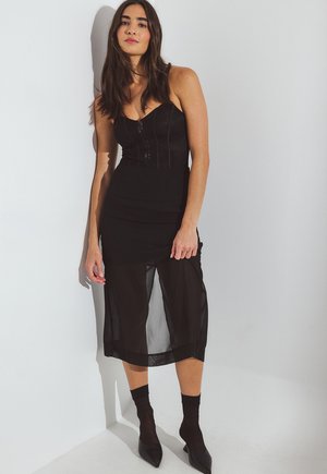 Kvinde der står op mod en hvid væg og har en sort korset-top, gennemsigtig midi-nederdel, sorte strømper og spidse sorte stilethæle på.