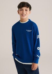 Modra teksturirana majica s dolgimi rokavi, okroglo izrezano in belim napisom "BLUE RIDGE DENIM". Vsebuje logotipne dodatke na rokavih.