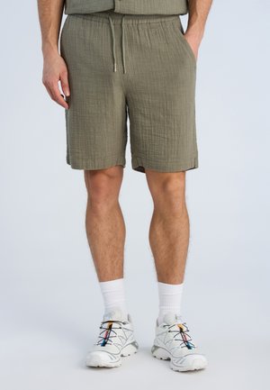 Man draagt olijfgroene gestructureerde korte broek met trekkoord, witte sokken en witte sportschoenen met kleurrijke strepen.