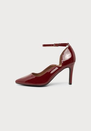 Chaussure à talon haut bordeaux brillante avec bout pointu et bride à la cheville sur fond gris clair.