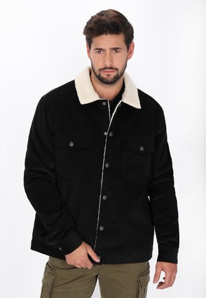 Bărbat cu barbă purtând o geacă neagră cu guler din fleece alb și pantaloni cargo verzi, stând în fața unui fundal alb.