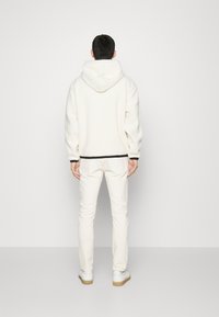 Weißer Fleece-Hoodie mit Kapuze, schwarzer Saumabschluss, kombiniert mit weißen Hosen und weißen Turnschuhen. Weiche Textur, lockere Passform, minimalistisches Design.