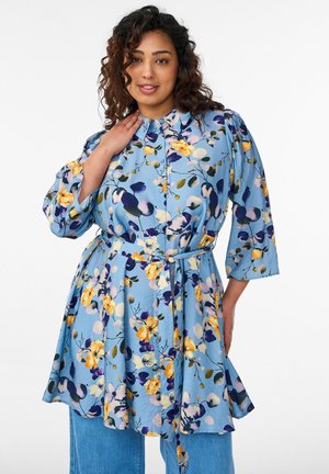 Žena s kučeravými vlasmi, ktorá nosí opásanú dlhú tuniku s kvetinovým vzorom v modrej, žltej a fialovej farbe, v spojení s modrými džínsami, pózujúca s rukou pri krku.