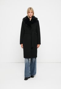 Cappotto nero con colletto in pelliccia, vita con cintura, maniche lunghe e tasche. Indossato con jeans in denim a gamba larga e stivali neri alla caviglia.