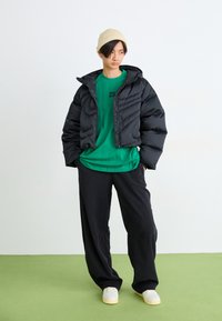 Casaco acolchoado preto com capuz, t-shirt verde com um gráfico, calças largas pretas, gorro creme e ténis brancos sobre um chão verde.