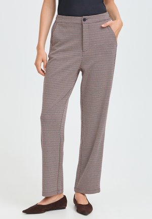 Femme portant un pantalon à carreaux taille haute, coupe droite, avec des poches latérales et des ballerines pointues marron foncé, debout devant un fond uni.