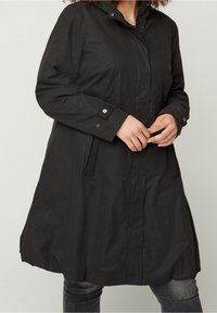 Cappotto nero fino al ginocchio con cappuccio, dotato di chiusura a zip, dettagli con bottoni a pressione e tasche laterali. Tessuto morbido e leggero.