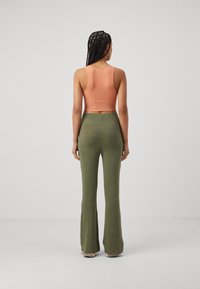 Femme aux cheveux tressés, debout de dos, portant un crop top pêche sans manches et un pantalon évasé taille haute vert olive.
