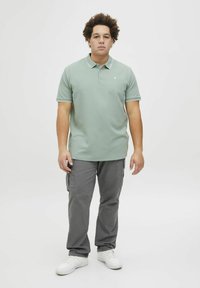 Lys grøn polo-shirt med et struktureret mønster og hvid kant, kombineret med grå cargo-bukser og hvide sneakers. Modellen står foran en ensfarvet baggrund.