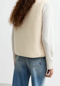 Person with long curly hair wearing a cream-colored textured vest over a white sweater and light blue faded jeans, shown from the back.