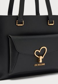 Τσάντα tote από μαύρο δερματίνη με χρυσές λεπτομέρειες, με κλειδαριά σε σχήμα καρδιάς και τσέπη με καπάκι στο μπροστινό μέρος. Το λογότυπο γράφει "LOVE MOSCHINO".