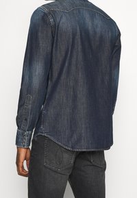 Granatowa koszula dżinsowa z długimi rękawami, spiczastym kołnierzykiem i mankietami zapinanymi na guziki. Posiada zaokrąglony dół oraz detale w kontrastowym przeszyciu.