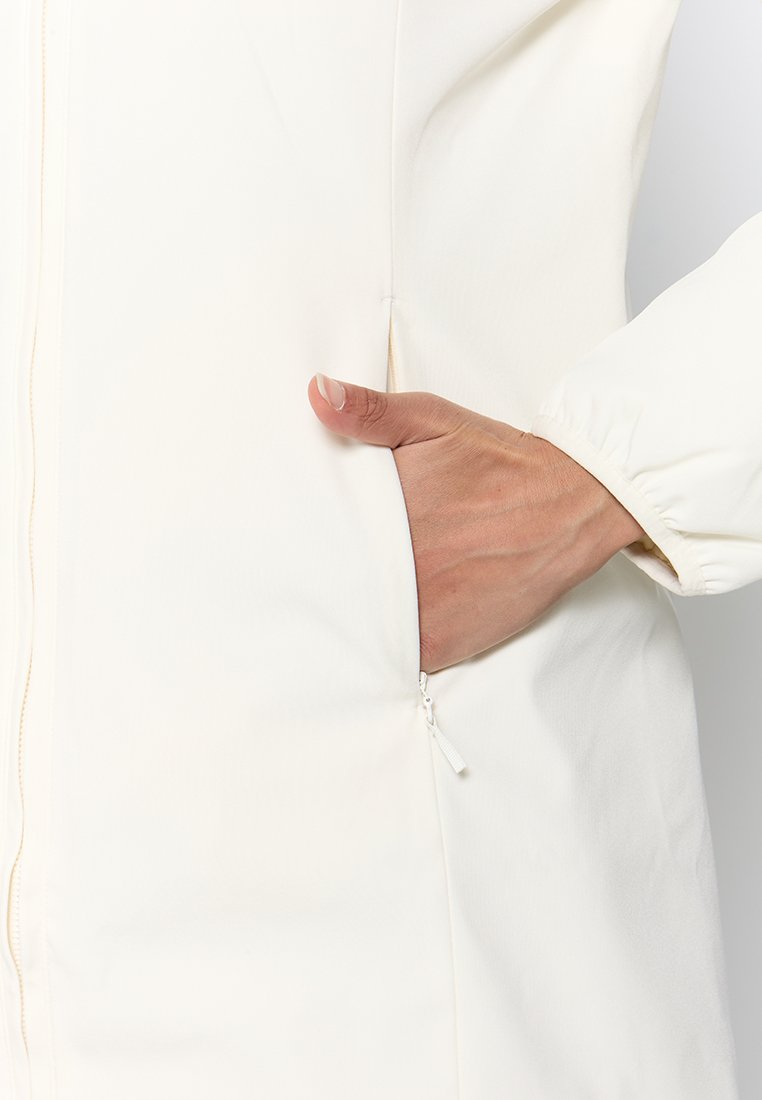 Cappotto lungo bianco con una texture liscia, dotato di una tasca laterale e un dettaglio con zip. Il design include un polsino aderente e un primo piano di una mano.