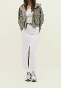 Vrouw in een cropped olijfgroen jasje, witte geribbelde cropped top, hoog in de taille gesneden witte midi-rok met een split aan de voorkant, en zilveren sneakers.