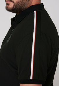 Czarna koszulka polo z kołnierzykiem na trzy guziki, z czerwonymi i białymi paskami na ramionach oraz emblematem na klatce piersiowej.