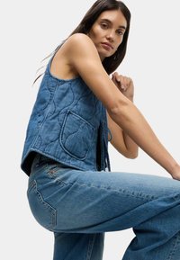 Jeansväst utan ärmar med vadderad textur, med en stor ficka och blå nyans, kombinerad med vida blå jeans.