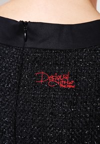 Svart texturerad tyg med en liten synlig dragkedja och röd broderad "Desigual" logotyp på den övre bakre midjebandet.