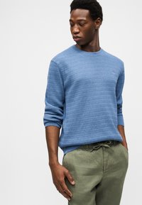 Νεαρός άνδρας που φοράει ένα μπλε πουλόβερ με υφή και ελιάς πράσινο παντελόνι, κοιτάζοντας προς τα κάτω αριστερά με το ένα χέρι στην τσέπη του.