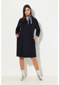 Marineblaues, knielanges Kleid mit Dreiviertelärmeln, Taschen und V-Ausschnitt. Kombiniert mit metallic-silbernen Ankle Boots und einem gemusterten Schal.