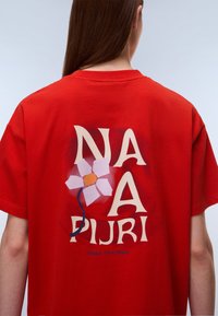 Κόκκινο βαμβακερό μπλουζάκι με μεγάλο γραφικό στην πλάτη που γράφει "NA PURI", σχέδιο λευκού λουλουδιού και το κείμενο "Make Your Mark" με μπλε χρώμα.