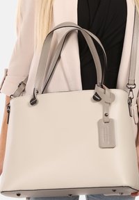 Beige lederen handtas met dubbele handvatten, een gevlochten accent en een afneembare tag. Voorzien van zilverkleurige hardware en een gestructureerde vorm.