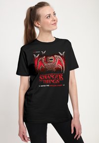 Camiseta negra con un diseño gráfico de un monstruo, el logo de "Stranger Things" y acentos de arte pixelado en rojo y blanco. Corte casual.