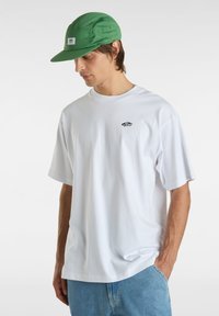 Weißes Baumwoll-T-Shirt mit kurzen Ärmeln und einem kleinen schwarzen Logo auf der Brust, kombiniert mit einer grünen Mesh-Cap mit einem Aufnäher auf der Vorderseite.