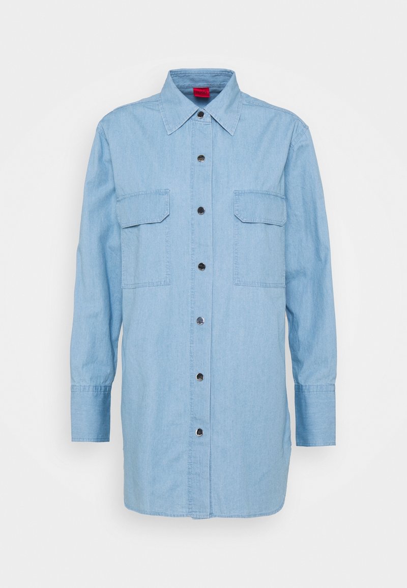 Chemise longue en denim bleu clair à manches longues, avec col, boutons métalliques sur le devant, et deux poches à rabat sur la poitrine.