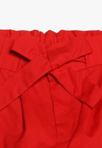 Czerwony materiał o gładkiej teksturze, z marszczoną talią i dużą kokardą z przodu, z widocznymi akcentami ściegów.