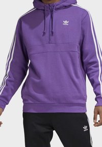 Lila hoodie med halvdragkedja, med vita ränder på ärmarna och en broderad logotyp på bröstet. Mjuk materialkänsla.