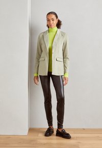 Ljusgrå blazer med snedskärning över en ljusgrön polotröja, kombinerat med blanka svarta leggings och svarta loafers.