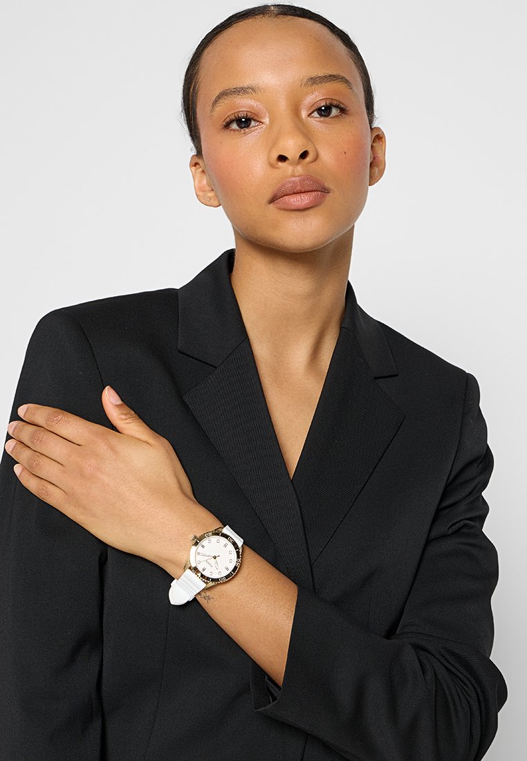Mujer con el cabello recogido que lleva una chaqueta negra, con los brazos cruzados, mostrando un reloj de pulsera con correa blanca y bisel dorado en su muñeca izquierda.