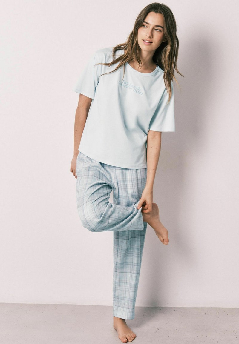 Femme en haut bleu clair à manches courtes et pantalon de pyjama à carreaux, debout pieds nus, tenant un ourlet de pantalon contre un mur clair.