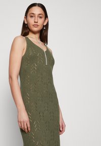 Robe tricotée sans manches couleur olive, présentant un motif texturé de lignes verticales et de motifs en forme de flèche. Longueur mi-mollet avec une silhouette ajustée.