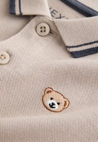 Beige textured polo shirt with two beige buttons, navy-striped collar, and embroidered teddy bear face on the chest.