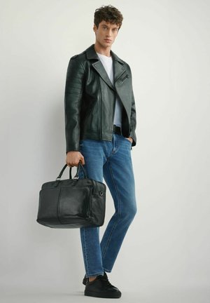 Młody mężczyzna w czarnej skórzanej kurtce, białej koszuli, niebieskich dżinsach i czarnych trampkach, trzymający czarną skórzaną aktówkę.