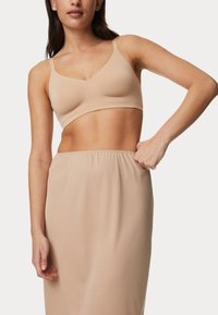 Sujeta beis sin costuras con tirantes ajustables, textura suave y forma natural. La modelo lleva una falda elástica a juego de color beis.