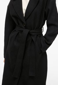 Svart beltefrakk med knappelukking, båret over en lys topp, med den ene hånden i lommen, mot en ensfarget lys bakgrunn.